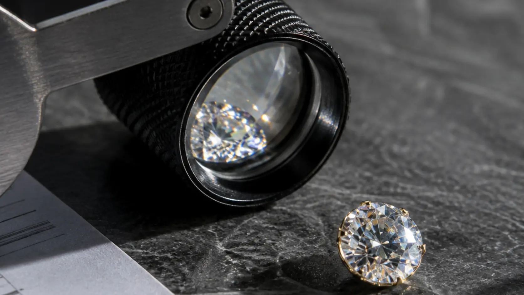 Lupa de joyero profesional junto a diamante suelto brillante sobre superficie de pizarra gris oscura con documento financiero parcialmente visible, representando el análisis técnico y financiero de las 4C para elegir el diamante perfecto sin pagar de más