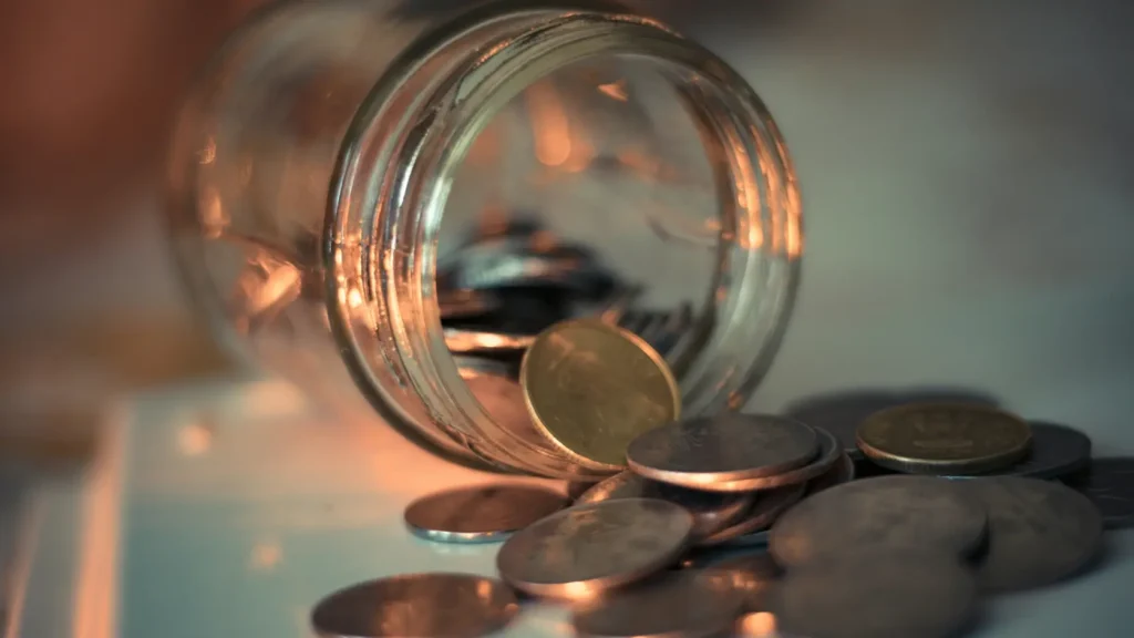 Tarro de cristal con monedas y billetes sobre mesa de cocina doméstica con luz natural, representando el plan de ahorro mensual para pagar la luna de miel sin financiación ni crédito antes de la boda